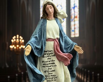 May include: A statue of the Virgin Mary, depicted with a serene expression, wearing a white dress, a blue cloak, and a white head covering. The statue has a pink sash and text on the front. The background shows a church interior with stained glass windows.
