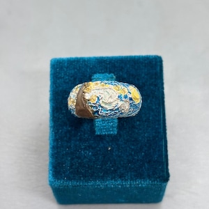 Può includere: Un anello dettagliato con un design blu, giallo e bianco, posto su un portagioie in velluto blu. L'anello presenta un motivo dettagliato con una fascia marrone. L'anello è un gioiello unico.
