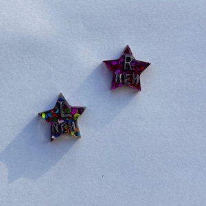 May include: Two star-shaped pins with glittery surfaces. One pin is a mix of colors, the other is a deep pink. Both pins have the letters "R NEH" printed on them. The pins are likely for decorative purposes.