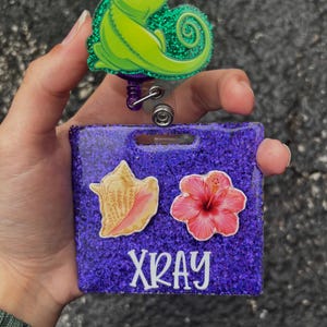 May include: A purple glitter badge reel with a cartoon green chameleon and the word "XRAY". The badge reel has a seashell and a pink hibiscus flower design.
