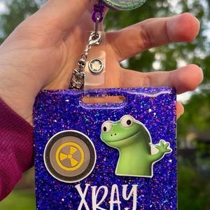 May include: A purple glitter badge holder with a cartoon frog and the word "XRAY" in white. The holder also features a radiation symbol and a skeleton charm. The retractable reel has a mermaid scale design.