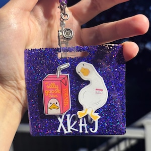 May include: A purple glitter badge holder with a cartoon goose and juice box design. The juice box reads "Silly Goose Juice" and the goose has the word "Silly" on it. The badge holder has the letters "XKAY" on it.