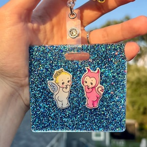May include: A square badge reel featuring a blue glitter background with two cartoon figures: a white angel with a halo and wings, and a pink devil with horns and a tail. Above, a gold glitter topper displays the text GOOSE BUMPS in black letters.