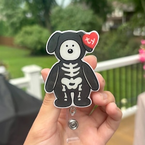 May include: A black and white badge reel featuring a cartoon dog character with a skeleton print on its torso. The dog has a white face, black ears, and a red heart-shaped tag near its ear with the letters ty printed in white on the surface.