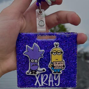 May include: A purple glitter badge holder with cartoon characters and the word "XRAY" in white. The holder features a purple cartoon character and a yellow Minion holding a sign that says "I'M WITH STUPID". A retractable reel with a Mike Wazowski charm is attached.