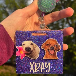 May include: A purple glitter badge reel with two dog photos and the word "XRAY" in white. The reel has a mermaid-themed top with a red bow and a green scale design. The badge reel is held by a person.