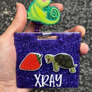 May include: A purple glitter badge holder with a green chameleon, a red strawberry, a green turtle, and the word "XRAY" in white. The badge holder has a retractable reel.