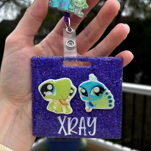 May include: A purple glitter badge holder with cartoon animal graphics and the word "XRAY" in white. The badge holder has a retractable reel with a cartoon monster design.