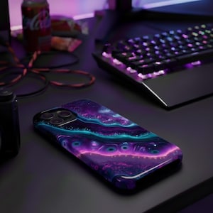 May include: A smartphone case with a vibrant, abstract design in shades of purple, blue, and pink. The case is lying on a black surface next to a Canon camera, a can of Coca-Cola, and a backlit keyboard.