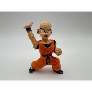 May include: A bald, light-skinned action figure in a dynamic martial arts pose. The figure wears a bright orange gi with a black undershirt, a black belt, dark wristbands, and dark shoes. The character has a determined facial expression.