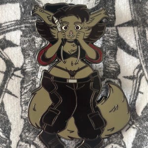 May include: Enamel pin featuring a stylized anthropomorphic wolf character. The pin has a dark color palette with black pants, boots, and a black top. The character has a knife and is holding its face. The pin has a silver outline and red accents.