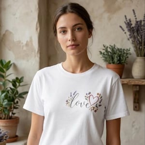 Peut inclure: T-shirt blanc avec le mot "love" en écriture cursive, accompagné d'un cœur et d'un motif floral. Le t-shirt à manches courtes a un col rond. Le motif comprend de la lavande et d'autres éléments floraux.