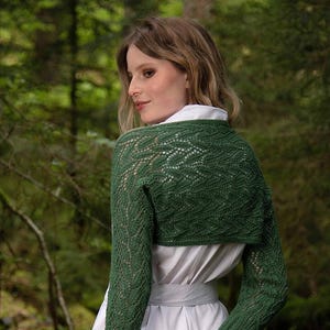 May include: A green knitted bolero with intricate openwork patterns. The bolero has long sleeves and is worn over a white dress. The background is a blurred view of a forest.