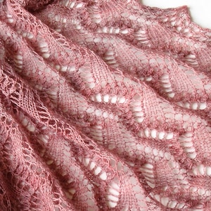 Könnte beinhalten: Nahaufnahme eines altrosa gestrickten Schals. Der Schal hat ein detailliertes Lochmuster mit einem gewellten Rand. Die Textur ist weich und das Muster erzeugt einen fließenden, wellenartigen Effekt. Der Schal ist wahrscheinlich aus einem zarten Garn gefertigt.