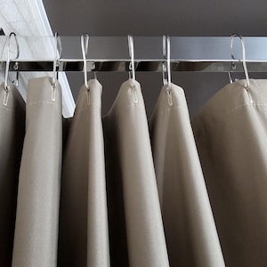 Puede incluir: Una cortina de ducha de color beige s&oacute;lido cuelga de una barra rectangular de cromo pulido. La cortina est&aacute; suspendida por ganchos en forma de S de metal plateado espaciados uniformemente a lo largo del borde superior, creando pliegues verticales.