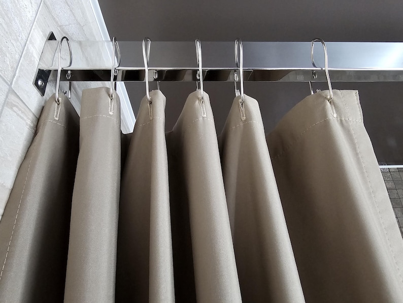 Puede incluir: Una cortina de ducha de color beige s&oacute;lido cuelga de una barra rectangular de cromo pulido. La cortina est&aacute; suspendida por ganchos en forma de S de metal plateado espaciados uniformemente a lo largo del borde superior, creando pliegues verticales.