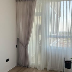 May include: Sheer, light-colored curtains in a modern room. One panel is a soft gray, tied back with a matching fabric tie. The other panel is white, allowing sunlight to filter through. The curtains frame a window with a view.