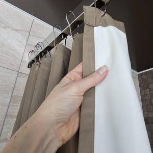 Puede incluir: Una cortina de ducha con un exterior de color topo s&oacute;lido y un forro interior blanco. La cortina est&aacute; suspendida de una barra de metal plateado mediante varios ganchos met&aacute;licos a juego, creando pliegues verticales en la tela.
