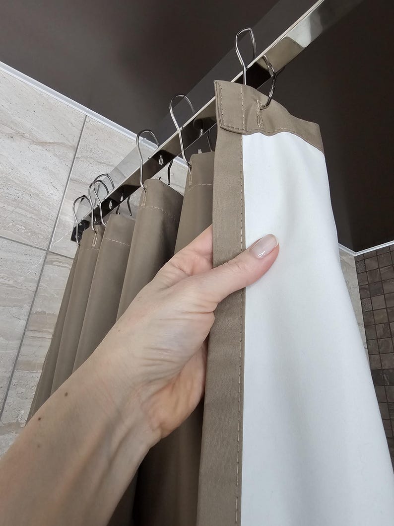 Puede incluir: Una cortina de ducha con un exterior de color topo s&oacute;lido y un forro interior blanco. La cortina est&aacute; suspendida de una barra de metal plateado mediante varios ganchos met&aacute;licos a juego, creando pliegues verticales en la tela.