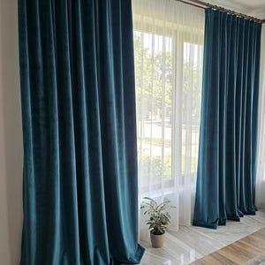 Puede incluir: Cortinas de terciopelo verde azulado colgadas de una barra, enmarcando una ventana con cortinas blancas transparentes. Las cortinas pesadas tienen pliegues profundos, creando una apariencia lujosa. Una pequeña planta en maceta está en el alféizar.