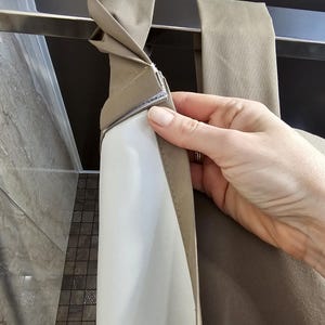 Puede incluir: Una cortina de ducha de tela color topo con un borde superior que presenta bucles de tela integrados que se pliegan sobre una barra met&aacute;lica horizontal. La cortina incluye un forro interior blanco unido a la capa exterior con una tira de velcro.