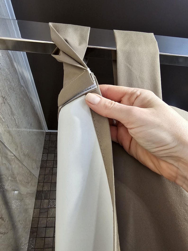 Puede incluir: Una cortina de ducha de tela color topo con un borde superior que presenta bucles de tela integrados que se pliegan sobre una barra met&aacute;lica horizontal. La cortina incluye un forro interior blanco unido a la capa exterior con una tira de velcro.