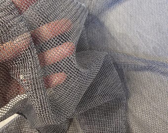 Puede incluir: Primer plano de una tela de malla plateada con un patrón tejido. La tela está drapeada y parece ligera. La malla se sostiene en una mano, mostrando la textura y transparencia del material.