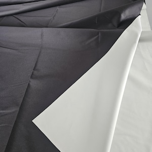 Puede incluir: Una pieza de tela con una capa superior de color gris carb&oacute;n oscuro doblada hacia atr&aacute;s para revelar un reverso liso de color blanco s&oacute;lido. El material presenta un acabado mate con pliegues y arrugas visibles en la superficie, mostrando un contraste claro entre los lados oscuro y claro del textil.