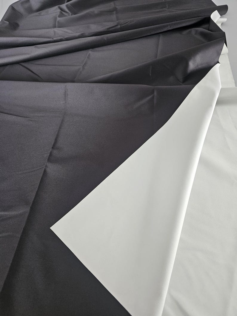 Puede incluir: Una pieza de tela con una capa superior de color gris carb&oacute;n oscuro doblada hacia atr&aacute;s para revelar un reverso liso de color blanco s&oacute;lido. El material presenta un acabado mate con pliegues y arrugas visibles en la superficie, mostrando un contraste claro entre los lados oscuro y claro del textil.