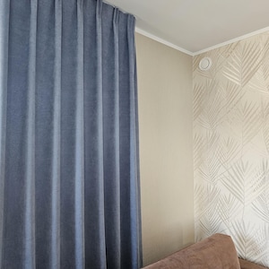 Puede incluir: Una cortina azul cuelga de un techo blanco, con pliegues verticales que crean una apariencia texturizada. La cortina está al lado de una pared con un papel pintado beige y blanco con estampado de hojas y un sofá marrón.