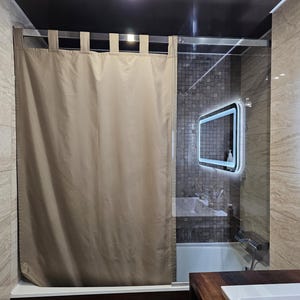 Puede incluir: Una cortina de ducha de color beige s&oacute;lido con un dise&ntilde;o de trabillas de tela que cuelgan de una barra de metal plateado. La cortina est&aacute; colocada frente a una mampara de ducha de cristal, ocultando parcialmente un ba&ntilde;o con azulejos.