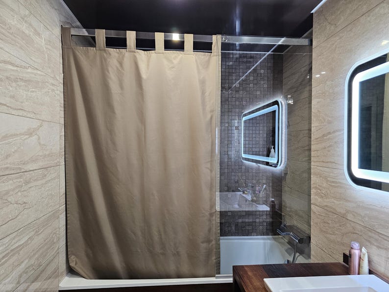 Puede incluir: Una cortina de ducha de color beige s&oacute;lido con un dise&ntilde;o de trabillas de tela que cuelgan de una barra de metal plateado. La cortina est&aacute; colocada frente a una mampara de ducha de cristal, ocultando parcialmente un ba&ntilde;o con azulejos.