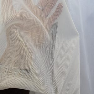 May include: Close-up of white, sheer fabric with a subtle herringbone pattern. The fabric is held up, allowing light to pass through, highlighting its delicate texture and weave. The material appears soft and lightweight.