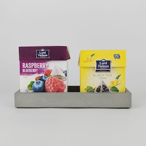 Op de afbeelding: Twee dozen Lord Nelson thee, één framboos-bosbes en één zwarte thee citroen, staan op een grijze betonnen tray. De framboos-bosbes doos is paars met fruit illustraties. De citroen thee doos is geel.