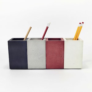 Peut inclure: Un organiseur de bureau rectangulaire avec quatre compartiments de couleurs différentes : bleu foncé, gris, rouge et blanc. Chaque compartiment contient un crayon ou un stylo. L'organisateur est en béton.