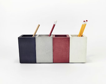 Op de afbeelding: Een rechthoekige bureau-organizer met vier compartimenten in verschillende kleuren: donkerblauw, grijs, rood en wit. Elk compartiment bevat een potlood of pen. De organizer is gemaakt van een betonachtig materiaal.