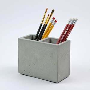 Puede incluir: Un organizador de escritorio de hormigón gris con dos compartimentos. Un lado contiene pinceles con mangos dorados y negros, el otro lado contiene lápices rojos con gomas blancas. El diseño es simple y moderno.