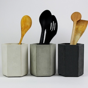 Può includere: Tre portautensili esagonali in cemento grigio chiaro, grigio medio e nero. Ogni contenitore contiene cucchiai di legno e utensili da cucina neri. Un elemento di arredo cucina moderno e minimalista.