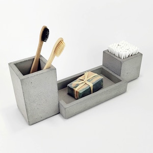 Può includere: Un set da bagno in cemento grigio con un portaspazzolini, una saponetta e un porta cotton fioc. Il portaspazzolini contiene due spazzolini. Una saponetta è nel portasapone, e i cotton fioc sono nel piccolo contenitore quadrato.
