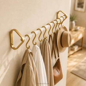 May include: A wall-mounted coat rack featuring a clear acrylic rod supported by two triangular gold-toned metal brackets. The rack includes several removable gold-toned S-shaped hooks holding a tan jacket, white shirt, scarf, bag, and hat.
