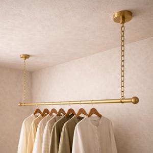Puede incluir: Un perchero dorado, montado en el techo, con una barra horizontal y una cadena colgante. Varias camisas de colores neutros cuelgan de perchas de madera. El perchero está suspendido del techo.