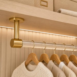 May include: A ceiling-mounted closet rod featuring a clear acrylic bar supported by brushed gold metal end caps and a central mounting bracket, with several wooden hangers holding various neutral-toned garments suspended from the rod.