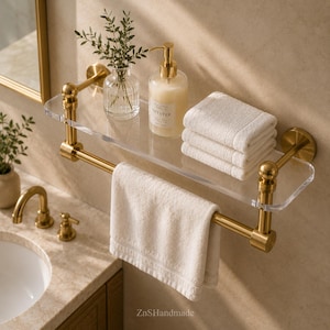 May include: A modern bathroom shelf with a clear acrylic surface and gold-colored metal accents. The shelf holds a stack of white towels, a soap dispenser, and a small vase with greenery. A white towel hangs from the lower bar.