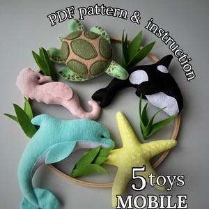 May include: A handmade mobile featuring felt sea creatures: a green turtle, a black and white orca, a pink seahorse, a blue dolphin, and a yellow starfish. The mobile includes the text "PDF pattern & instruction" and "5 toys MOBILE".