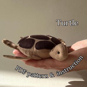 May include: A plush turtle toy in tan and brown, with the word "Turtle" above it. The turtle has a patterned shell and is held in a hand. Text below reads "PDF pattern & instruction".