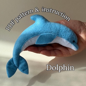 May include: A handmade blue and white felt dolphin toy. The dolphin is held in a hand, showcasing its curved body, fins, and tail. The text "PDF pattern & instruction" and "Dolphin" are visible.
