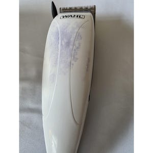 May include: A white Wahl hair clipper with a black blade guard. The clipper has a curved design and some purple discoloration on the body. The brand name "WAHL" is printed on the top.