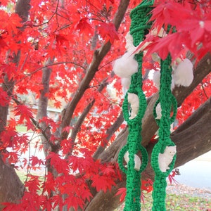 May include: A green and white macrame wall hanging with white pom-poms and a fringe. The hanging is in front of a tree with red leaves.