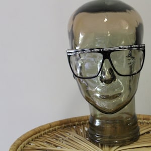 May include: A clear glass mannequin head wearing black framed glasses with a silver rhinestone trim. The glasses have a square shape and are sitting on a wicker surface.