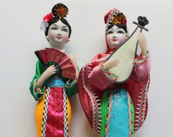 May include: Two traditional Chinese dolls wearing colorful silk dresses. One doll holds a fan, and the other plays a stringed instrument.
